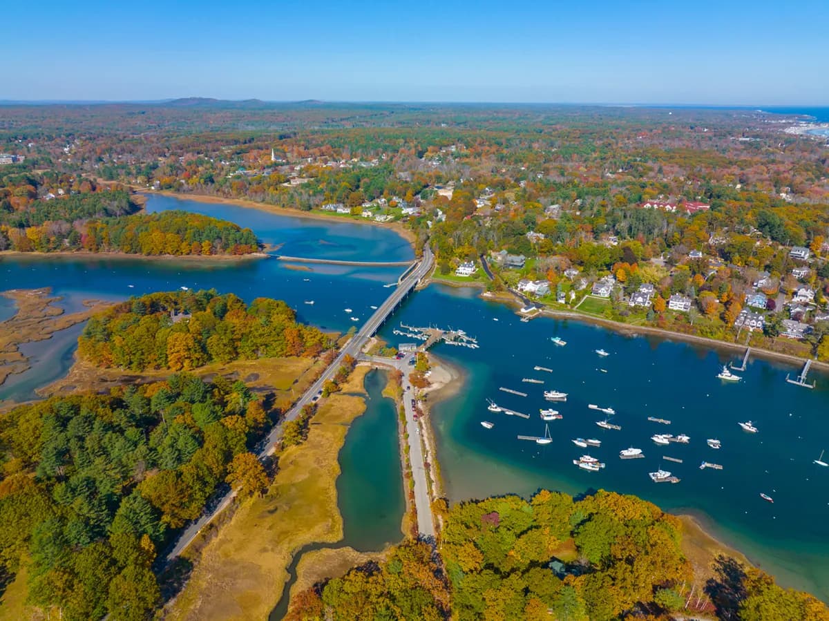 Aerial view of a coastal town in autumn with colorful trees, a bridge crossing blue water, and boats docked in a marina.