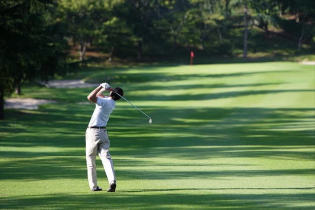 Golfer mid-swing on a lush green fairway surrounded by trees on a sunny day.
