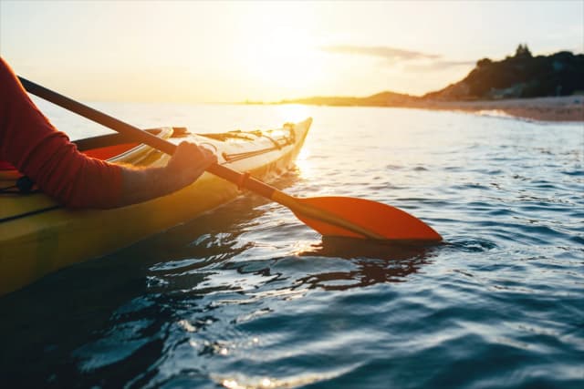 Person kayaking on calm ocean water at sunset, with warm sunlight reflecting off the waves.
