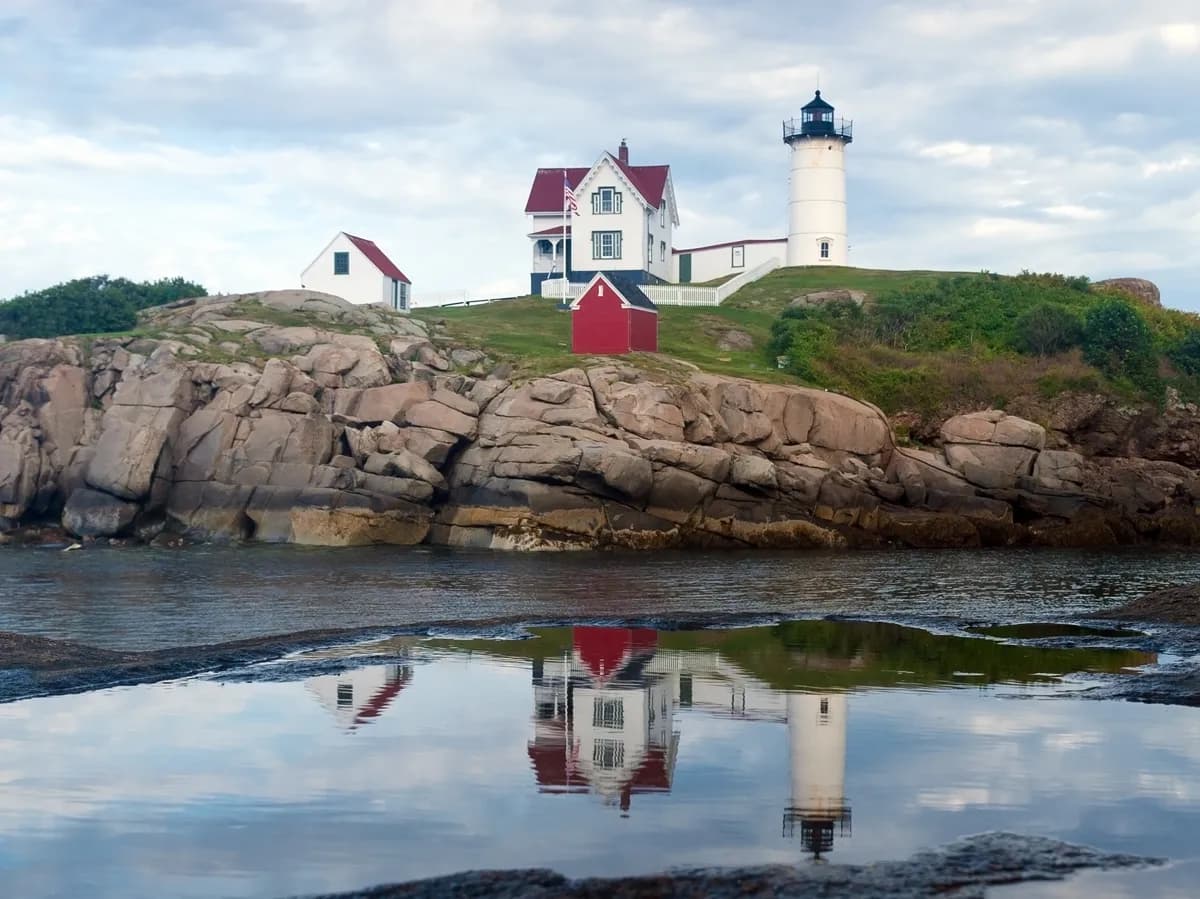 Coastal lighthouse and white seaside buildings on rocky cliffs with reflections in still water.