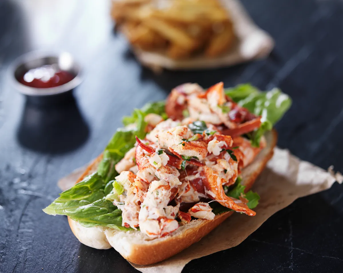 Fresh lobster roll served on a toasted bun with lettuce, accompanied by fries and a side of dipping sauce.